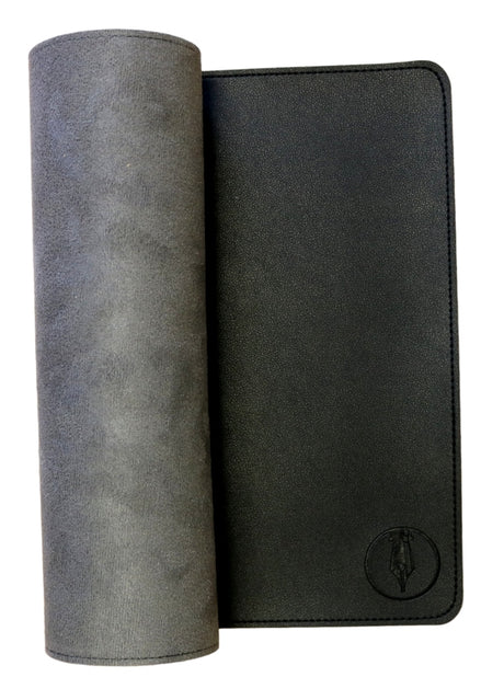 A black FPR Writing Mat, with one edge gently rolled up to reveal a soft interior evocative of a premium writing surface. The flat section showcases a small, embossed fountain pen logo in the lower right corner, mirroring the refined detailing characteristic of ink-resistant desk protectors.