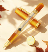 Nahvalur Original Plus Fountain Pen - Fountain Pen Day 2026 (Limited Edition) in clear Italian acrylic with a gold nib and metallic orange accents, shown with its cap and a matching circular Fountain Pen Day emblem.