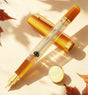 Nahvalur Original Plus Fountain Pen - Fountain Pen Day 2026 (Limited Edition) in clear Italian acrylic with a gold nib and metallic orange accents, shown with its cap and a matching circular Fountain Pen Day emblem.